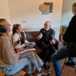 Singer/songwriter Heatherlyn and storyteller and guitarist Steve Bassett sit on a sofa and Rob Taylor sits on a stool in the foreground. Someone from The Hopeful Activist podcast sits on a stool and records an interview with them.