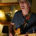 Steve Bassett looks to the right of the camera and smiles whilst playing an electric guitar.