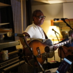 At rehearsals, Ben Okafor stands and smiles whilst holding his acoustic guitar. Heatherlyn laughs in the foreground and Steve Bassett sits and plays guitar in the background.