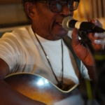 Ben Okafor sings into a microphone with his eyes closed. He holds the microphone with one hand and his acoustic guitar with the other.