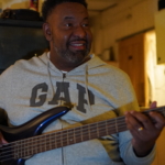 Tony Bean smiles and looks to his left whilst practising his five string bass guitar.