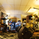 With amps in the foreground of the photo, the Nine Beats Collective stand or sit around a room and rehearse together.