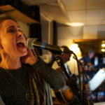 Heatherlyn sings passionately into a microphone. Ben Okafor, Steve Bassett and Tony Bean rehearse on guitars in the background.