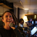 Heatherlyn stands in front of a microphone and laughs whilst looking to the left on the camera. Ben Okafor, Steve Bassett and Tony Bean rehearse on guitars in the background.