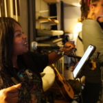 Belinda Kae smiles whilst singing into a microphone. Heatherlyn sings into a microphone in the background.