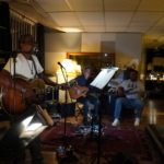 At rehearsals, Ben Okafor sings into a microphone and plays acoustic guitar. In the background, Steve Bassett and Tony Bean sit and play guitars too. To the right a drum kit is visible.