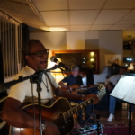 At rehearsals, Ben Okafor sings into a microphone and plays acoustic guitar. In the background, Steve Bassett and Tony Bean sit and play guitars too.