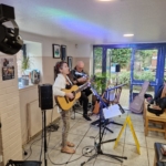 Singer/songwriter Heatherlyn stands and sings whilst playing guitar and guitarist Steve Bassett sits on a stool beside her playing an electric guitar. This rehearsal takes place during the day.