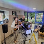 Singer/songwriter Heatherlyn stands and sings whilst playing guitar and guitarist Steve Bassett sits on a stool beside her playing an electric guitar. This rehearsal takes place during the day.