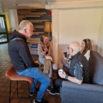 Looking from the left: Singer/songwriter Heatherlyn and storyteller and guitarist Steve Bassett sit on a sofa and Rob Taylor sits on a stool in the foreground. Someone from The Hopeful Activist podcast sits on a stool and records an interview with them.