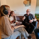 Singer/songwriter Heatherlyn and storyteller and guitarist Steve Bassett sit on a sofa whilst someone from The Hopeful Activist podcast sits on a stool and records an interview with them.