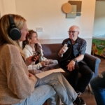 Singer/songwriter Heatherlyn and storyteller and guitarist Steve Bassett sit on a sofa whilst someone from The Hopeful Activist podcast sits on a stool and records an interview with them.