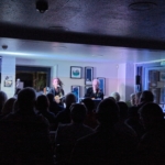 Singer/songwriter Heatherlyn stands and sings whilst playing guitar and guitarist Steve Bassett sits on a stool beside her playing an electric guitar. An audience sits and watches them perform. There is a banner to the left that reads 'The Barn Acoustic Nights'.