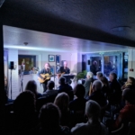 Singer/songwriter Heatherlyn stands and sings whilst playing guitar and guitarist Steve Bassett sits on a stool beside her playing an electric guitar. An audience sits and watches them perform.