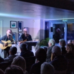 Singer/songwriter Heatherlyn stands and sings whilst playing guitar and guitarist Steve Bassett sits on a stool beside her playing an electric guitar. An audience sits and watches them perform.