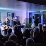 Singer/songwriter Heatherlyn stands and sings whilst playing guitar and guitarist Steve Bassett sits on a stool beside her playing an electric guitar. An audience sits and watches them perform.