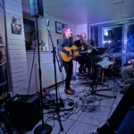 Singer/songwriter Heatherlyn stands and sings whilst playing guitar and guitarist Steve Bassett sits on a stool beside her playing an electric guitar.