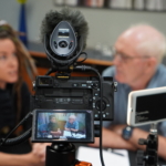 A camera records Heatherlyn and Steve Bassett as they speak during a virtual interview. Heatherlyn and Steve can be seen both on the camera's screen and out of focus in the background.