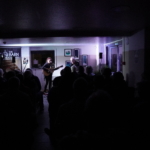 A dark photo taken from behind the audience. Singer/songwriter Heatherlyn stands and sings whilst playing guitar and guitarist Steve Bassett sits on a stool beside her playing an electric guitar. An audience sits and watches them perform.