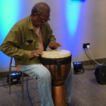 Ben Okafor sits and plays a drum that he holds between his knees.