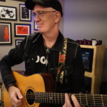 Steve Bassett smiles whilst playing an acoustic guitar.