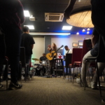 From the view of the floor of the aisle, Heatherlyn sings, whilst Tony Bean plays bass guitar and Marcus Copeland plays drums behind her.