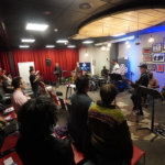 Keyboardist Martin Trotman, lead guitarist Rich Watson, drummer Marcus Copeland, singer/songwriter Heatherlyn, reggae artist Ben Okafor and guitarist Steve Bassett perform in front of an audience. There is a red curtain along the left wall and a grey wall with framed art behind the collective. The photo was taken from the right side of the audience.