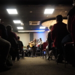 From the view of the centre of the aisle at the back, Heatherlyn sings and plays guitar. In the background Tony Bean plays bass guitar and Marcus Copeland plays drums. Members of the audience are visible on either side.