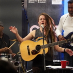 Heatherlyn sings into a microphone with both hands raised forwards and her eyes closed whilst holding an acoustic guitar. In the background Marcus Copeland plays the drums and Tony Bean plays a bass guitar.