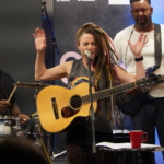 Heatherlyn sings into a microphone with both hands raised and her eyes closed whilst holding an acoustic guitar. In the background Marcus Copeland plays the drums and Tony Bean plays a bass guitar.