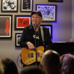 Steve Bassett smiles whilst speaking into a microphone and holding an electric guitar.