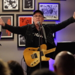 Steve Bassett performs spoken word with his hands raised, with an electric guitar.