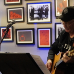 Steve Bassett plays acoustic guitar and reads the music on the stand in front of him with a gallery of framed pictures behind him.