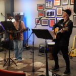 Ben Okafor sings whilst he and Steve Bassett play on acoustic guitars in front of a wall with a gallery of framed pictures. Tony Bean and Marcus Copeland are visible in the background.