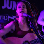 In front of a Record Junkee banner, Heatherlyn sings into a microphone whilst playing an acoustic guitar. Marcus Copeland and Tony Bean are slightly visible in the background.