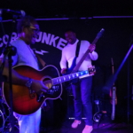 Ben Okafor and Tony Bean, both holding guitars, laugh together on the stage. To the side, Steve Bassett plays an acoustic guitar.