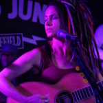 In front of a Record Junkee banner, Heatherlyn sings into a microphone with her eyes closed whilst playing an acoustic guitar. Marcus Copeland and Tony Bean are slightly visible in the background.