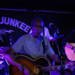 Tony Bean plays guitar in front of the Record Junkee banner. Steve Bassett, Marcus Copeland and Tony Bean play their instruments in the background too.