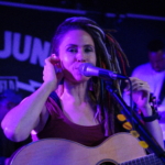 In front of a Record Junkee banner, Heatherlyn sings into a microphone with one hand raised to her ear whilst holding an acoustic guitar. Marcus Copeland and Tony Bean are slightly visible in the background.