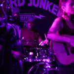 Marcus Copeland plays the drums at Record Junkee. Belinda Kae and Heatherlyn are visible out of focus in the foreground and Tony Bean is partially visible in the background.