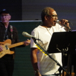 Ben Okafor performs a song whilst holding the microphone on the stand. Steve Bassett plays guitar in the background.