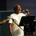 Ben Okafor performs a song whilst holding the microphone on the stand. Steve Bassett plays guitar in the background.