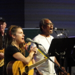 Ben Okafor and Heatherlyn sing into microphones whilst Steve Bassett plays guitar in the background.