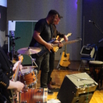 With a view from the right hand side of the band, Tony Bean and Steve Bassett, both with guitars, stand together laughing. Rich Wilson is partially visible to the left playing guitar, as is the drum kit, two acoustic guitars, two amps and a hand drum.