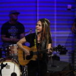 Heatherlyn sings into a microphone whilst playing guitar. Marcus Copeland plays drums in the background and Tony Bean and Ben Okafor are visible to the right hand side.