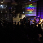 Ben Okafor plays a drum and the rest of the Nine Beats Collective performs to the audience with a Nine Beats Collective logos projected above the band. The silhouettes of the audience are visible in the foreground.