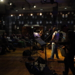 With a view from the left hand side of the visible, the audience is in view on the left in dark lighting and the band are in view on the right, with Steve Bassett playing guitar in the foreground.