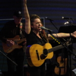 Heatherlyn sings passionately into a microphone with her left arm raised to the side and her right arm raised above, whilst holding an acoustic guitar. Steve Bassett plays guitar in the background.