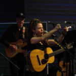 Heatherlyn sings passionately into a microphone with fists raised in the air whilst holding an acoustic guitar. Steve Bassett plays guitar in the background.