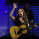 Heatherlyn sings into a microphone with a hand raised and her eyes closed whilst holding an acoustic guitar. Tony Bean plays a five string bass guitar in the background.